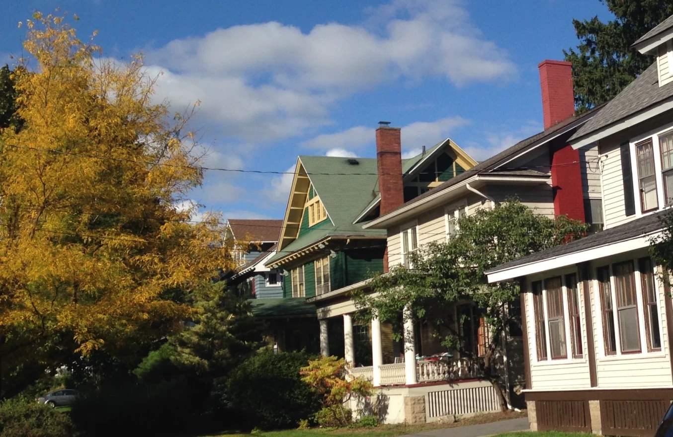 Three two story houses are seen side by side on a sunny day. The middle house has a large tree in the front yard with yellow leaves